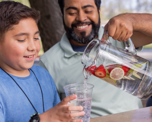 A man pouring fruit-infused water into a cup for a boy.