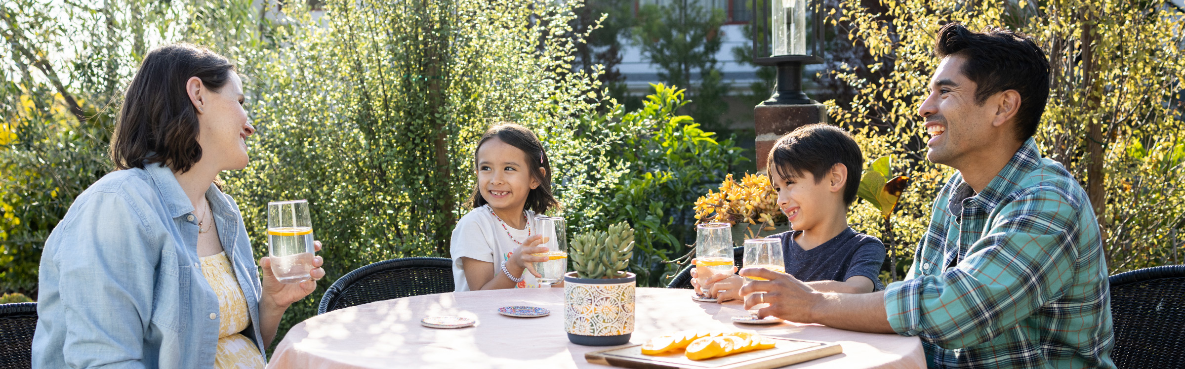 A family of four smiling while drinking fruit-infused water at a table in a sunny backyard.